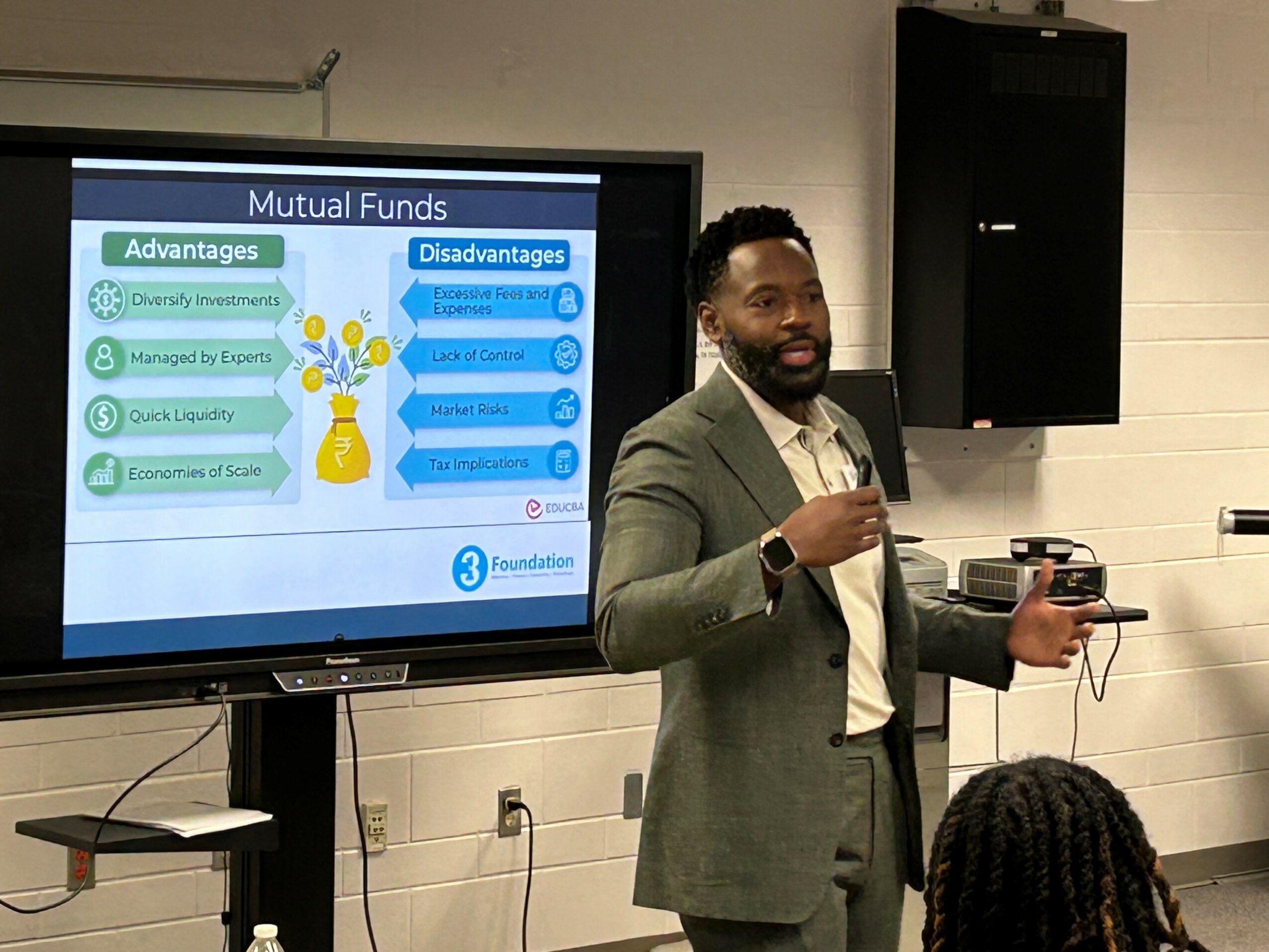 Man in gray suit presenting a slide on mutual funds to an audience in a conference room.