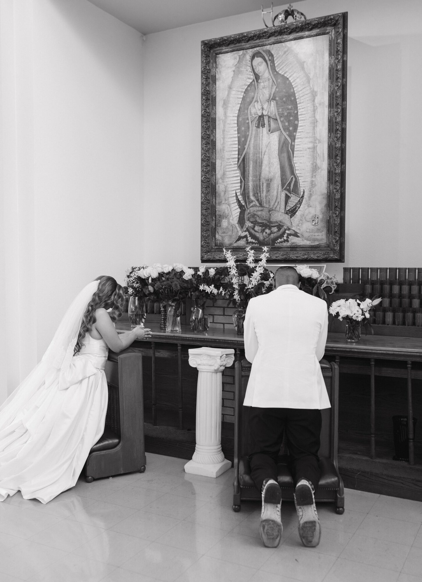 Bride and groom kneel at altar with flowers, religious painting hangs above, in a church setting.
