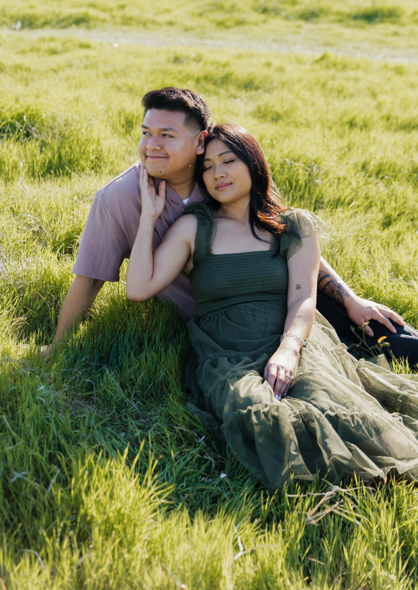 A man and woman sit on grass in a field, leaning against each other with relaxed expressions.