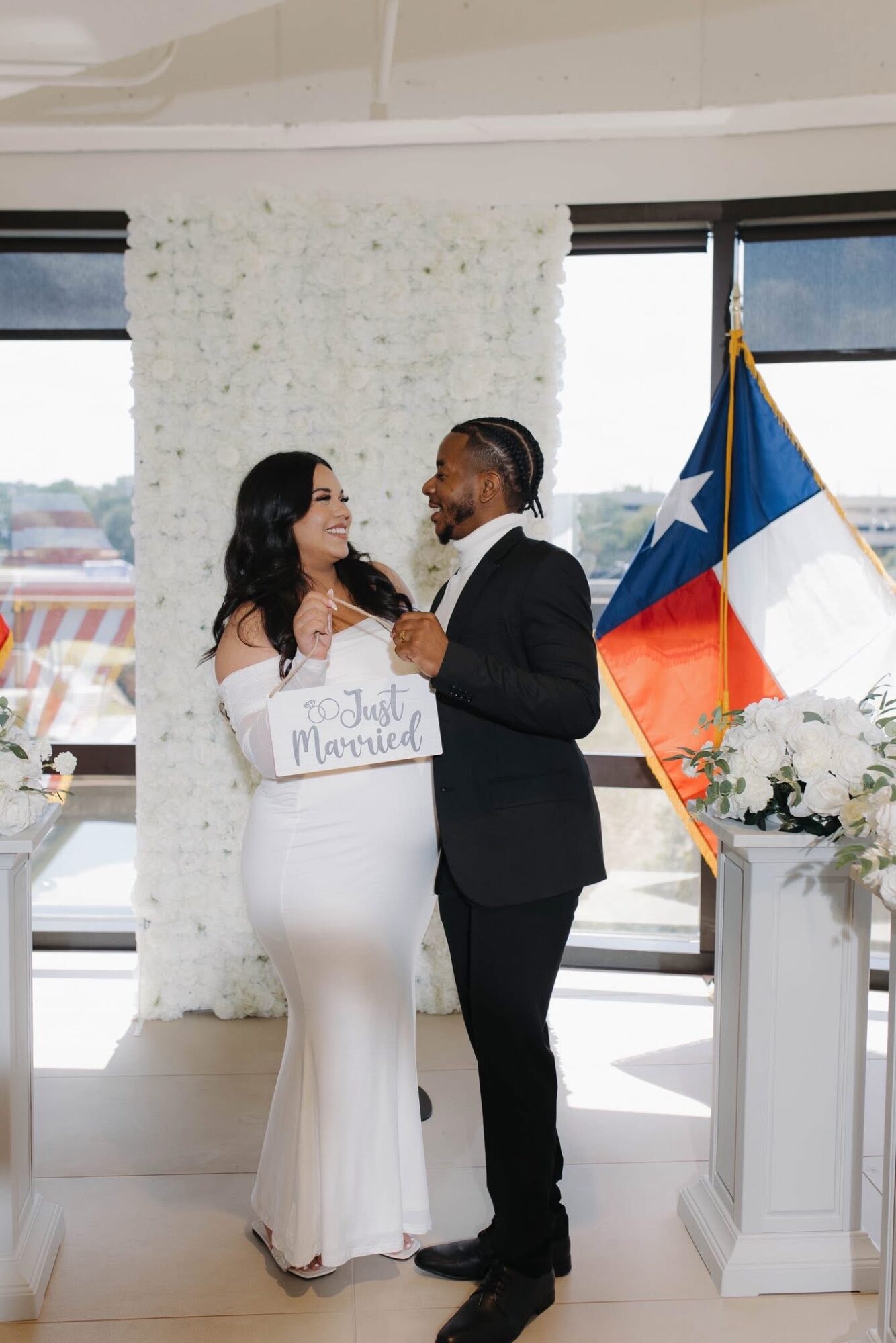 A couple stands indoors, holding a sign, with a Texas flag and flowers nearby, in front of large windows.