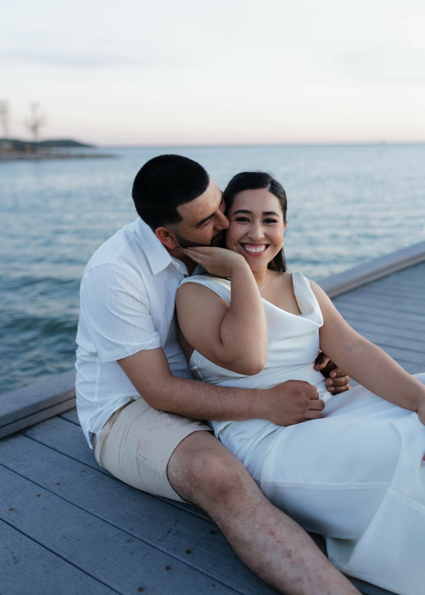 A couple sitting on a dock by the water, smiling and embracing each other.