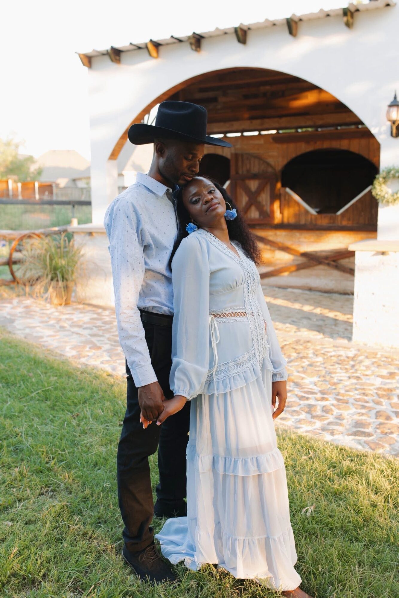 A couple stands outdoors near a white building with an arched opening, holding hands and leaning on each other.