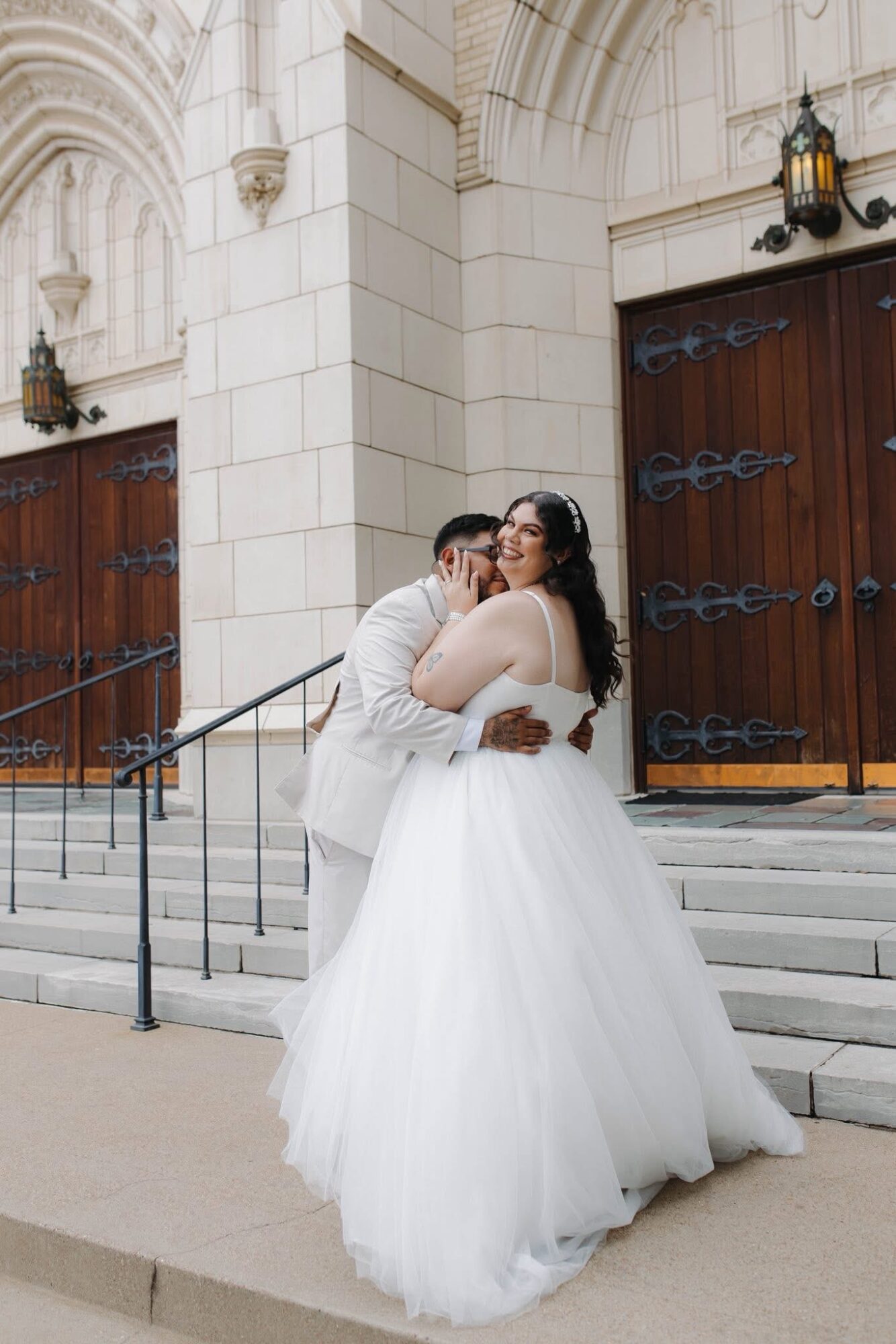Couple in wedding attire embracing on church steps, with large wooden doors and stone architecture in background.