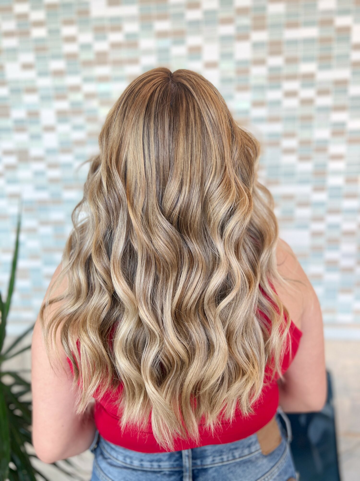Back view of a woman with long, wavy blonde hair wearing a red top and blue jeans.