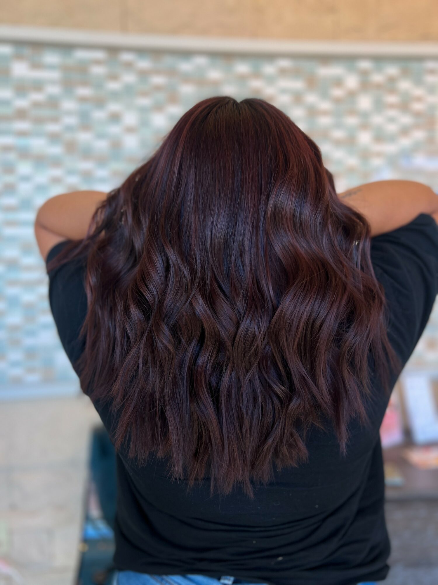 Back view of a person with long, wavy, dark brown hair, wearing a black shirt, near a tiled wall.