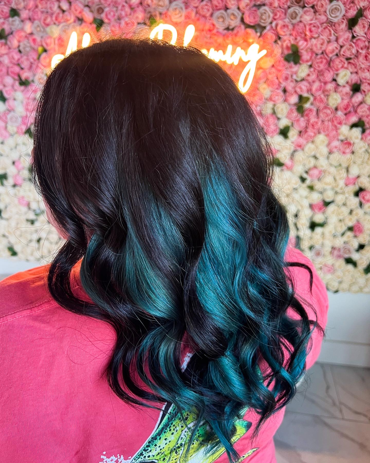 Back of a person with long, wavy black hair with blue highlights, wearing a pink top, in front of a floral wall.