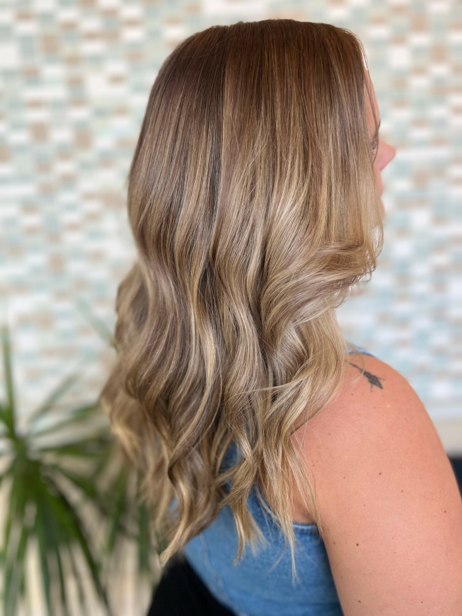 Side view of a woman with long, wavy, light brown hair with blonde highlights, wearing a sleeveless top.