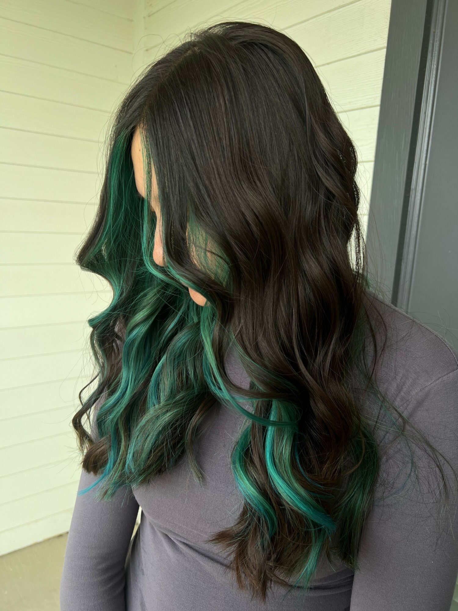 Person with long dark hair and green-tinted ends, standing outdoors near a wall and window.