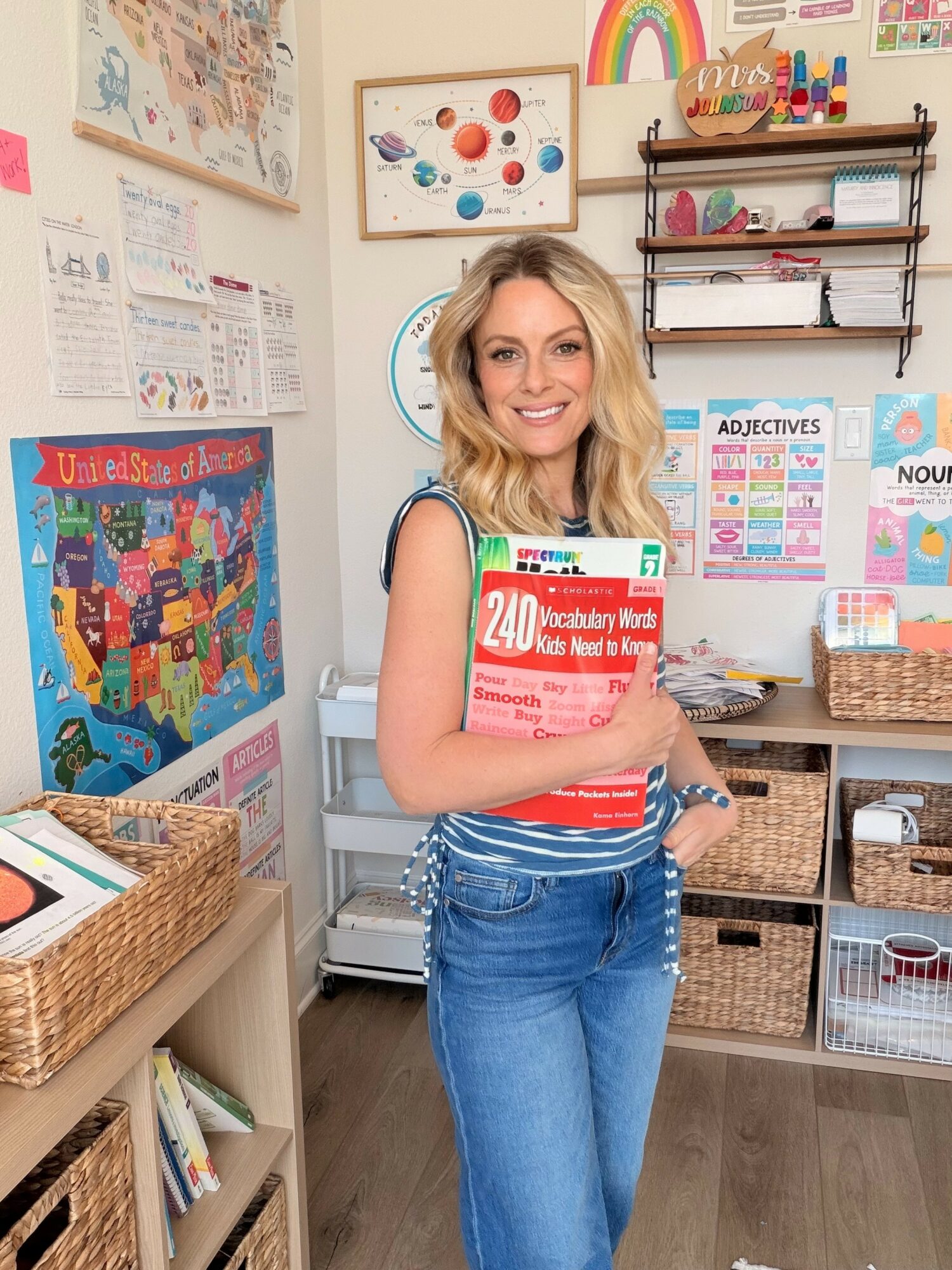 Woman with blonde hair holding a red book in a classroom with educational posters and shelves.