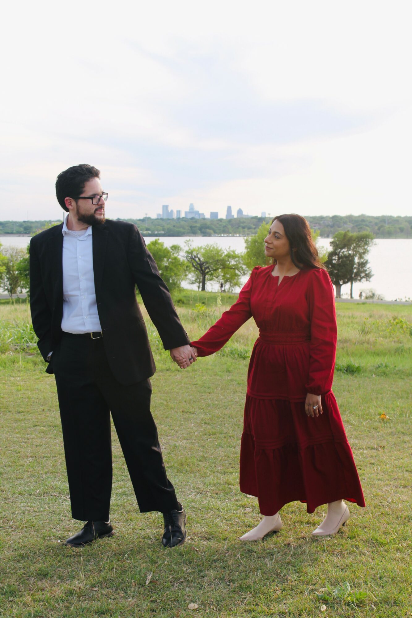 Man and woman holding hands in a grassy field with a city skyline in the background.