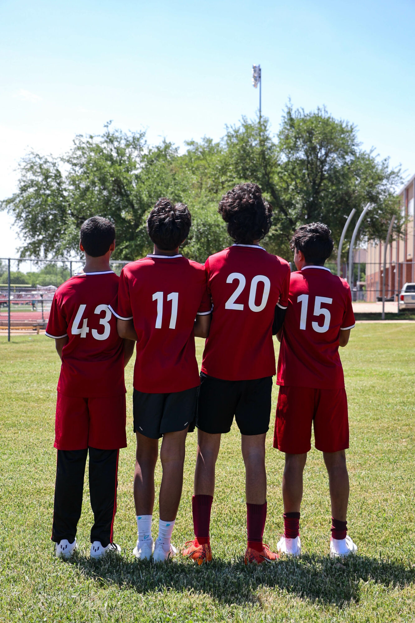 Four children in red sports uniforms standing on grass with their backs to the camera, facing a sports field.