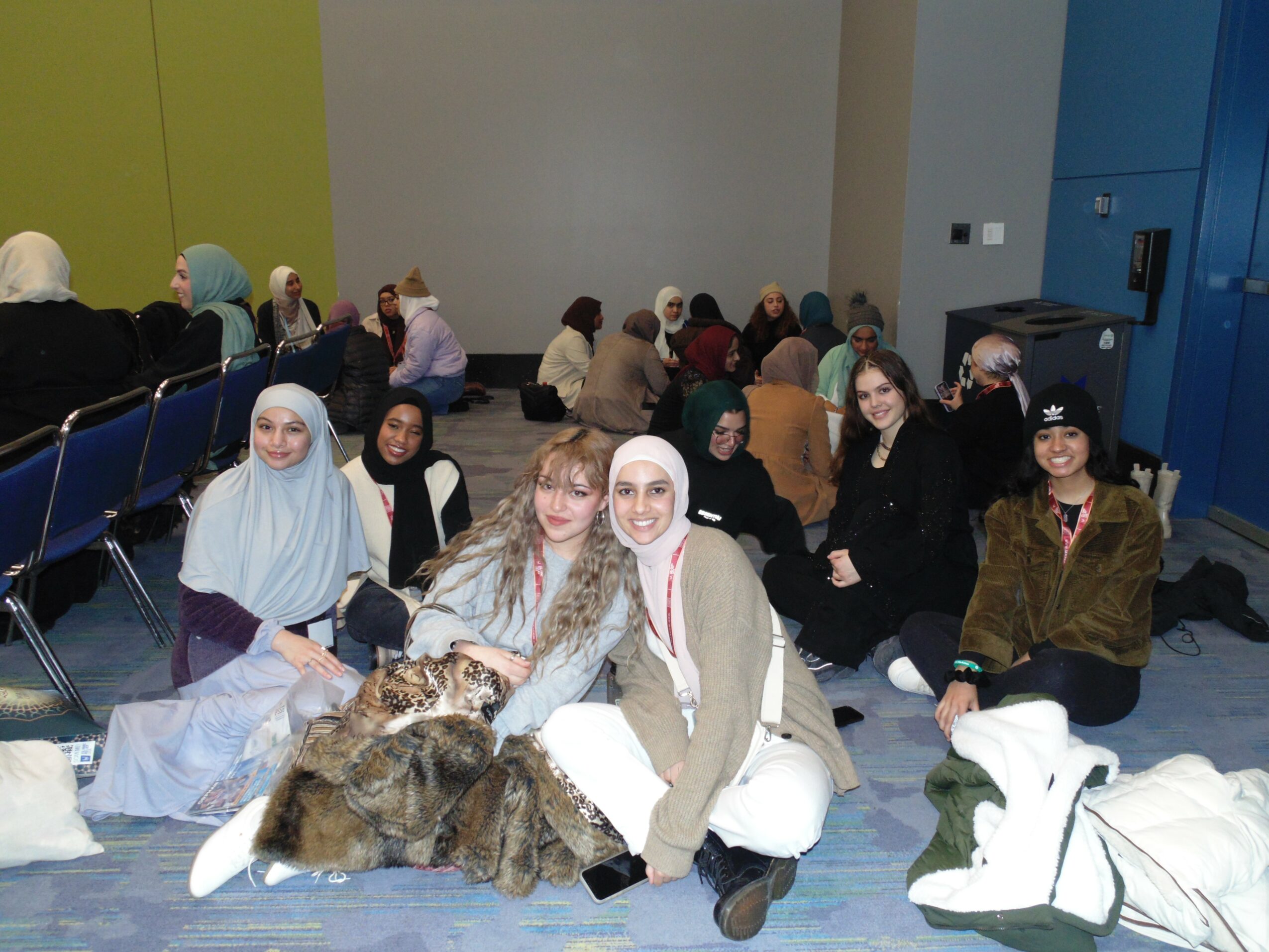 Group of women sitting on the floor and chairs in a room, some wearing headscarves, with bags and coats nearby.