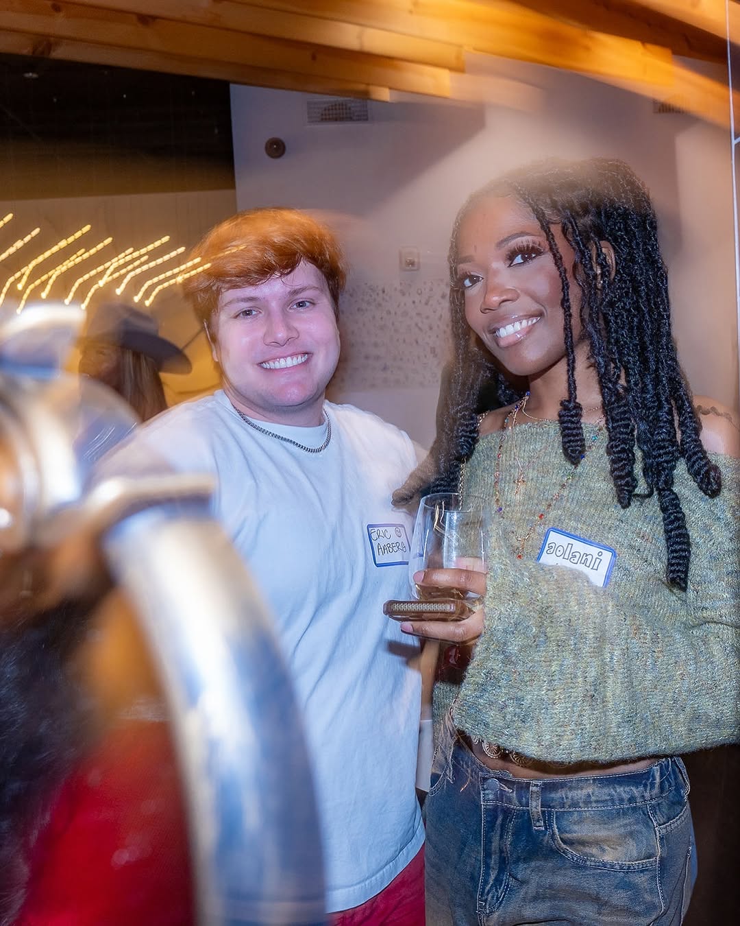 Two people smiling at a social event, one holding a glass, with warm lighting and blurred background.