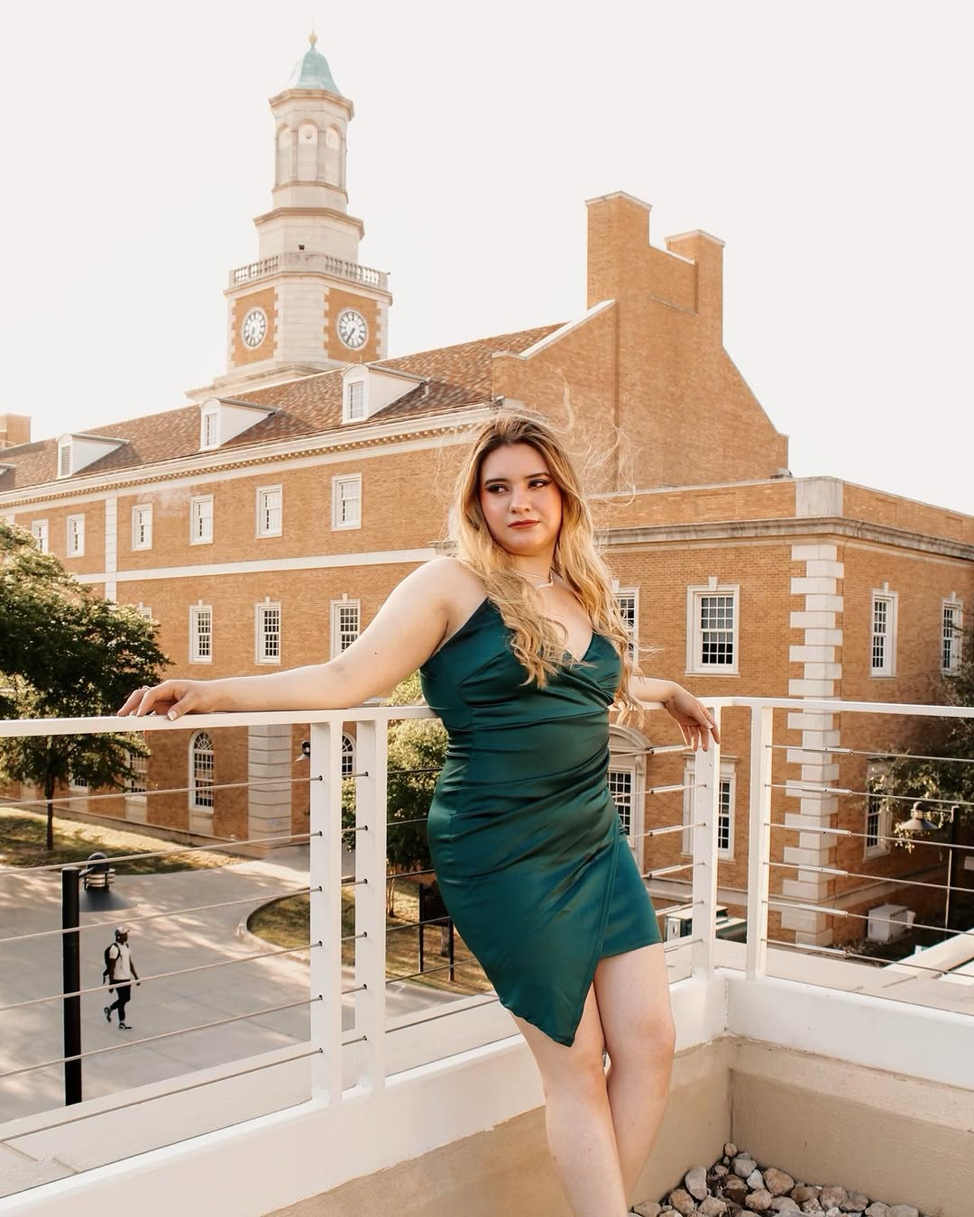 Woman in teal dress standing on balcony with historic brick building and clock tower in background.