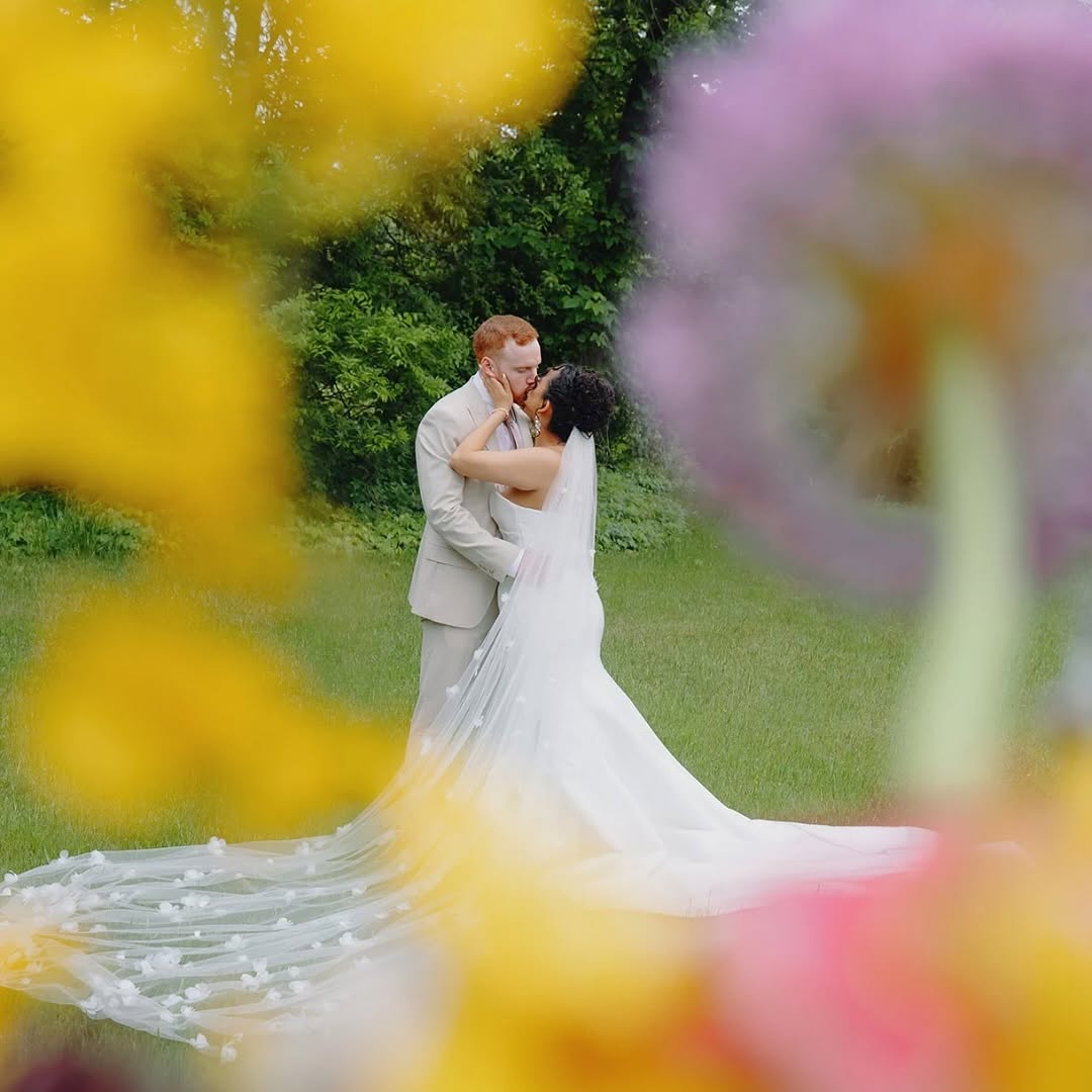 Bride and groom embrace outdoors, surrounded by colorful flowers and greenery, with blurred yellow and pink flowers in foreground.