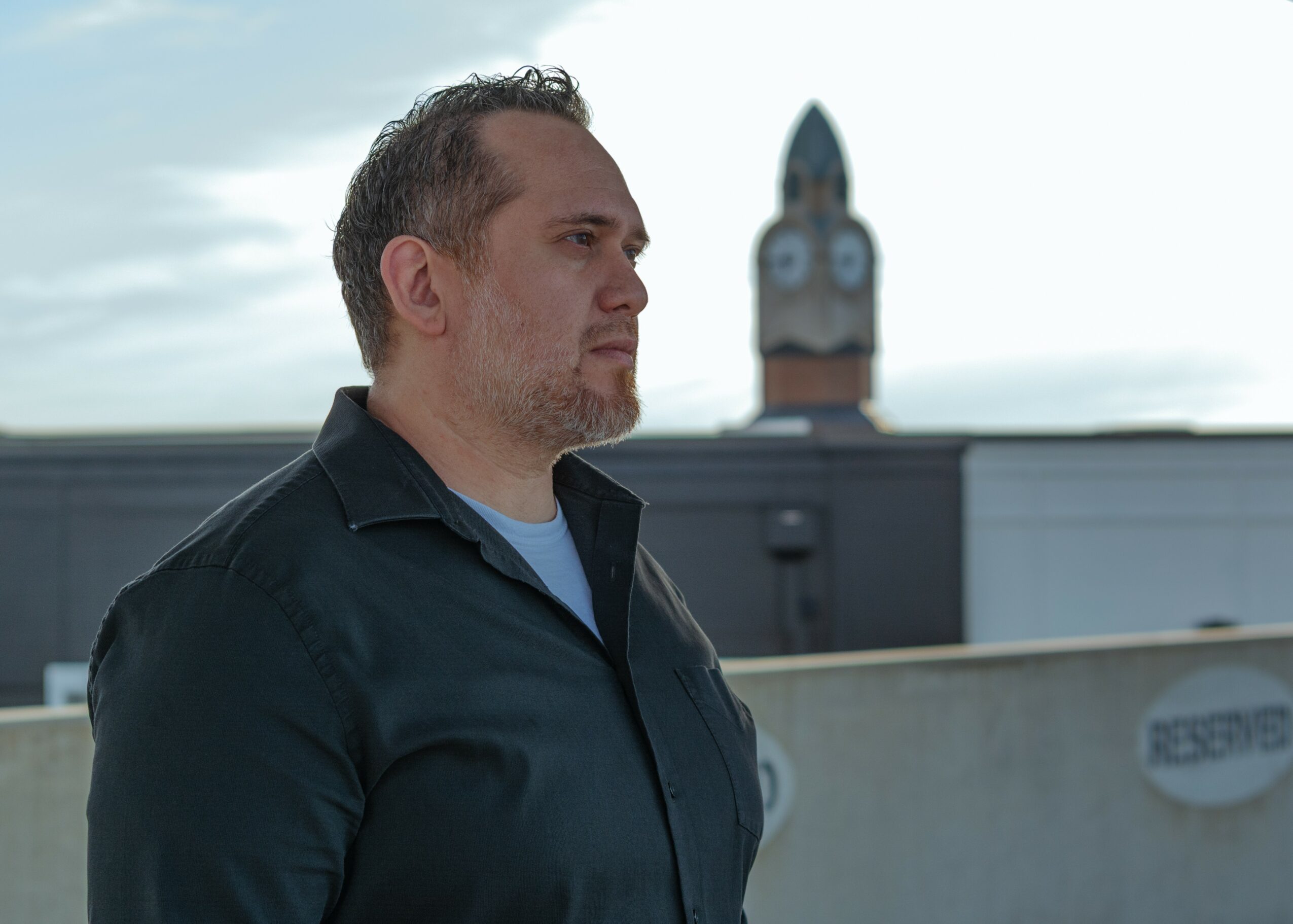 Man with short hair and beard in profile, standing outdoors with clock tower in background.