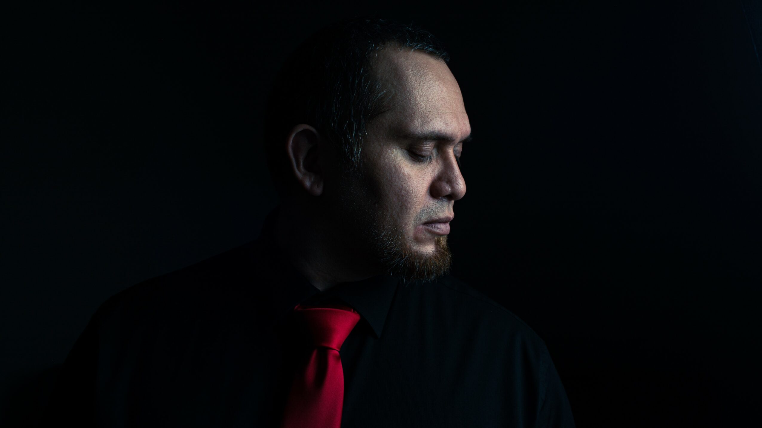 Side profile of a man with closed eyes, wearing a black shirt and red tie, against a dark background.