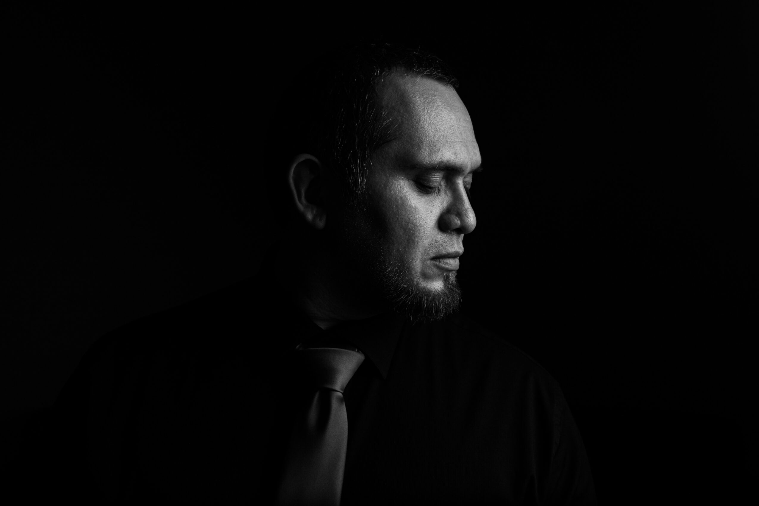 Man with closed eyes, wearing a suit and tie, facing left, in a dark background.