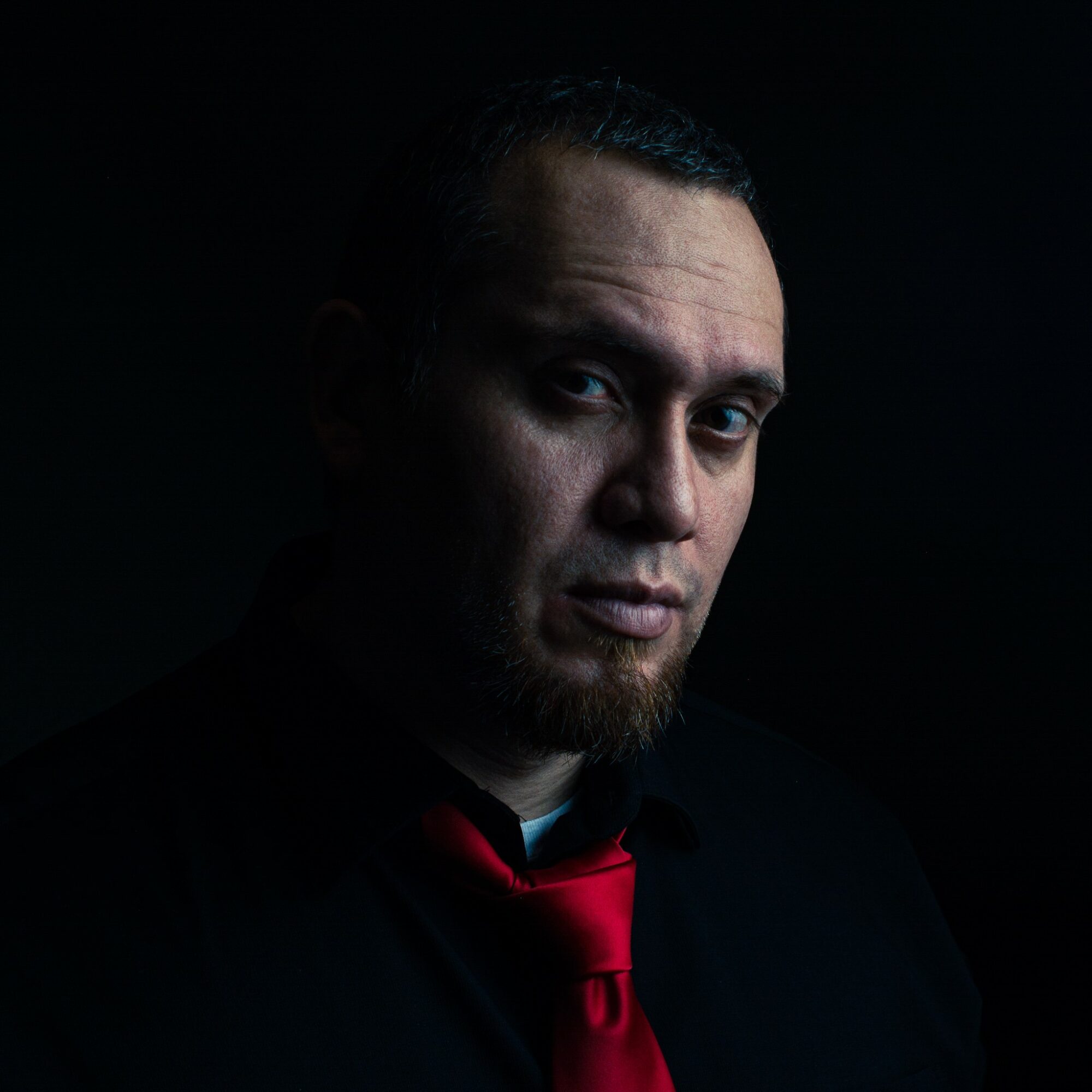 Portrait of a man with dark background, short hair, beard, wearing a black shirt and red tie.