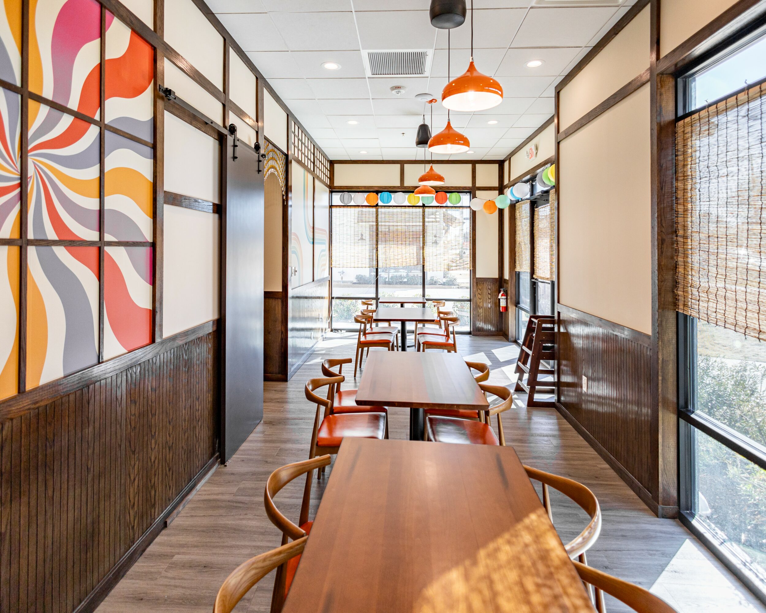 Empty restaurant with tables, chairs, colorful hanging lights, large windows, and patterned wall art.