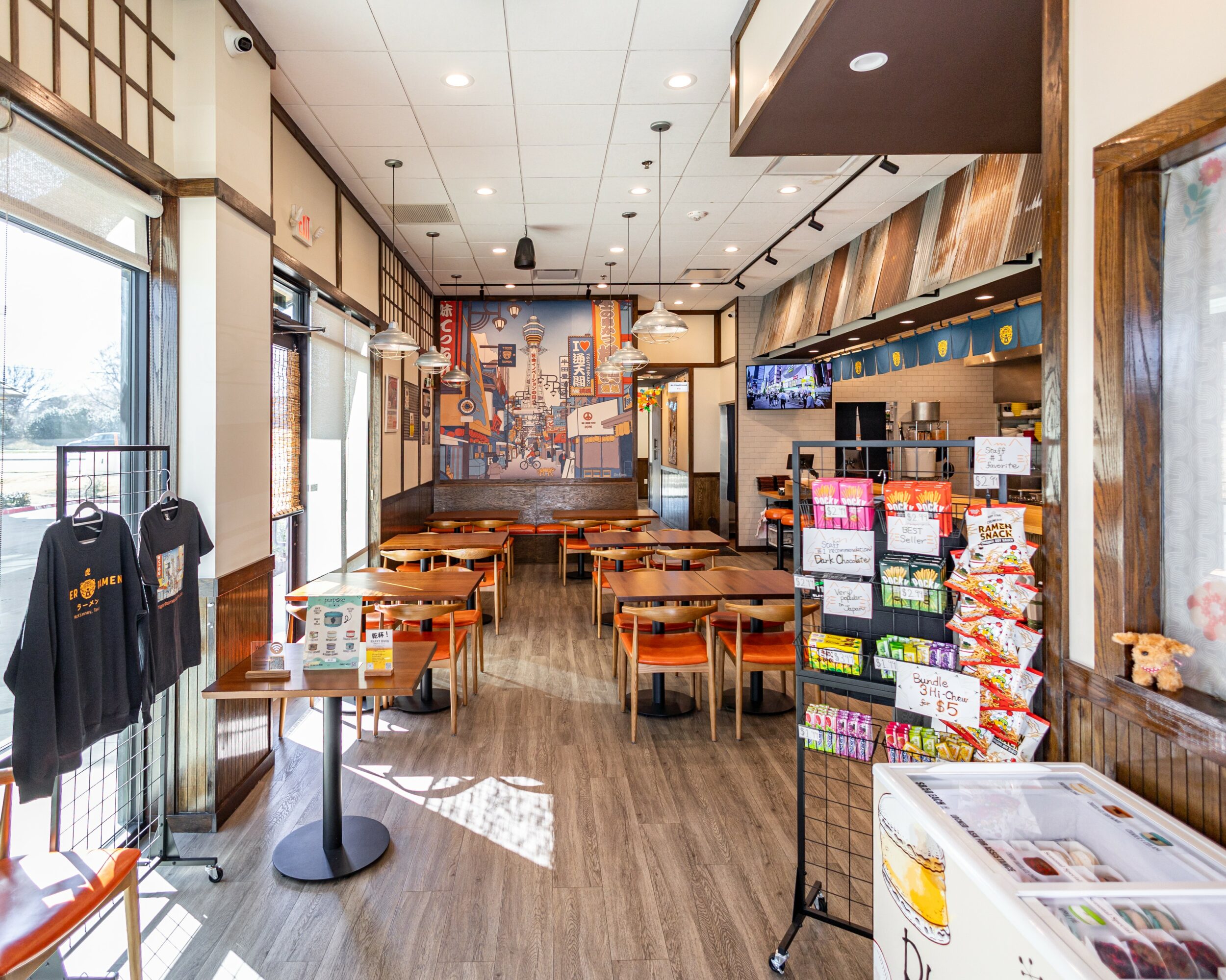 Empty restaurant with tables, chairs, and a display stand near the entrance, sunlight streaming through windows, and a colorful mural on the wall.