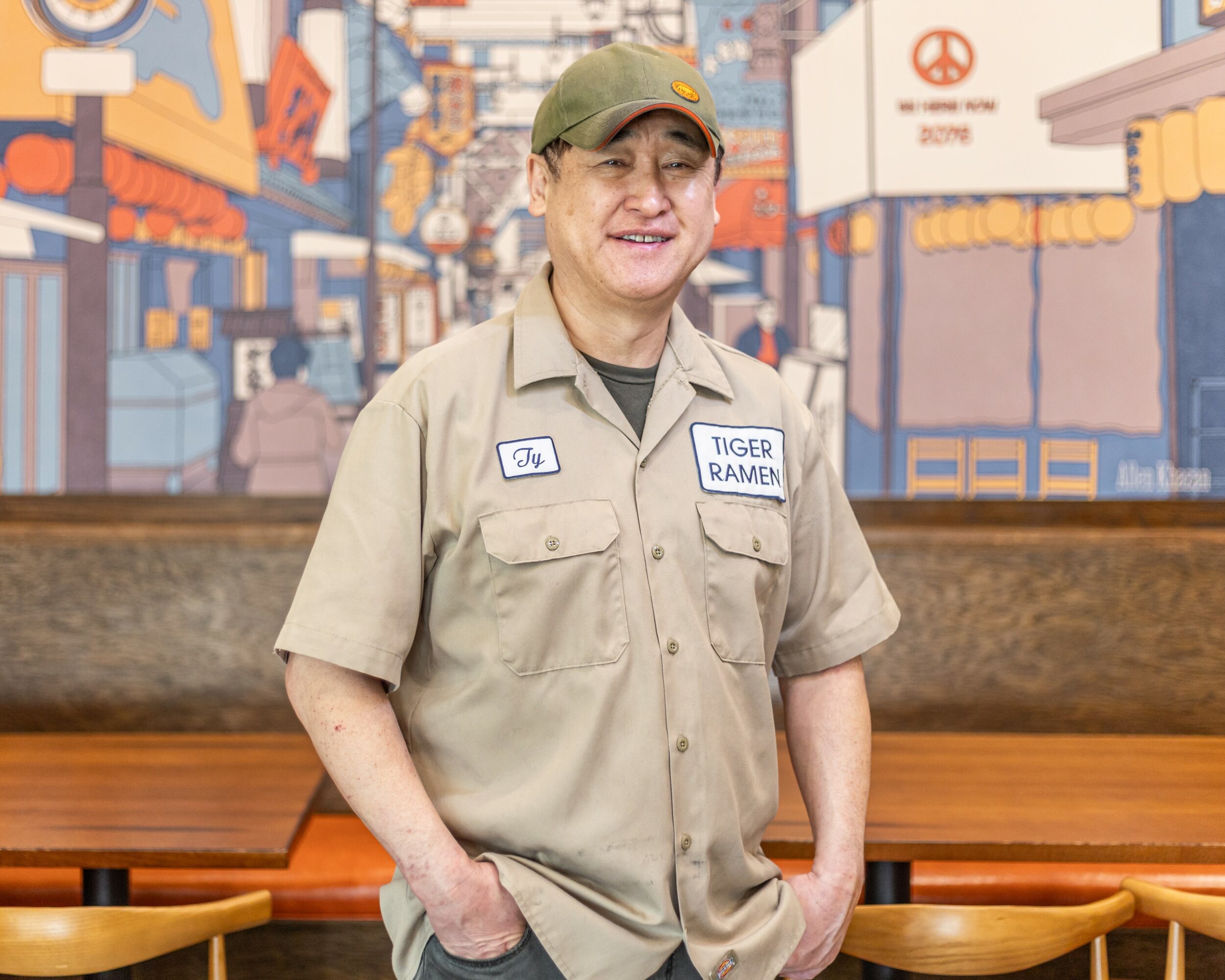 Smiling man in uniform standing indoors with colorful mural background, hands in pockets.