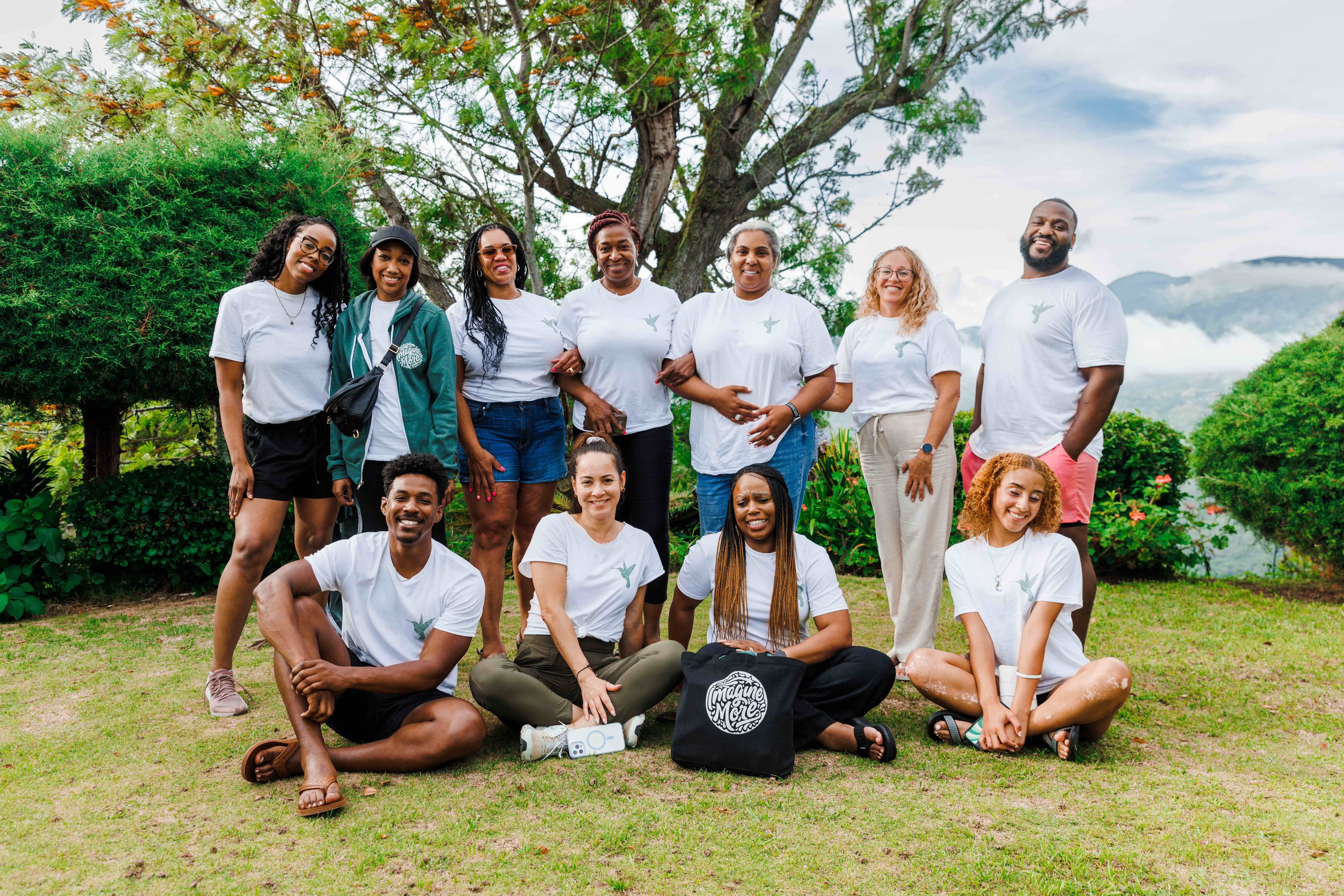 Group of ten diverse people outdoors, some standing and some sitting on grass, with trees and lake in background.