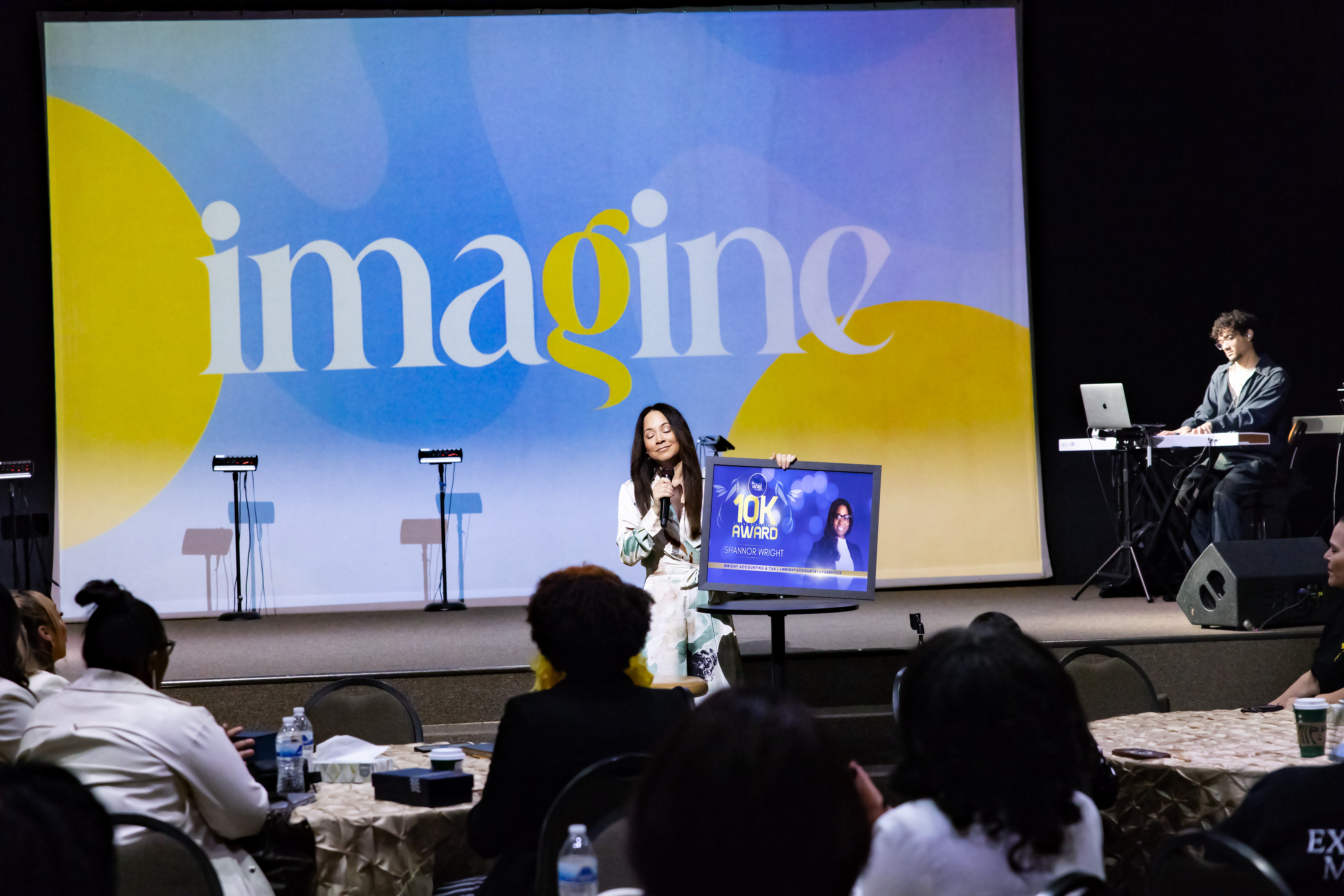 Person speaking on stage with large screen behind displaying the word 'imagine'. Audience seated watching.