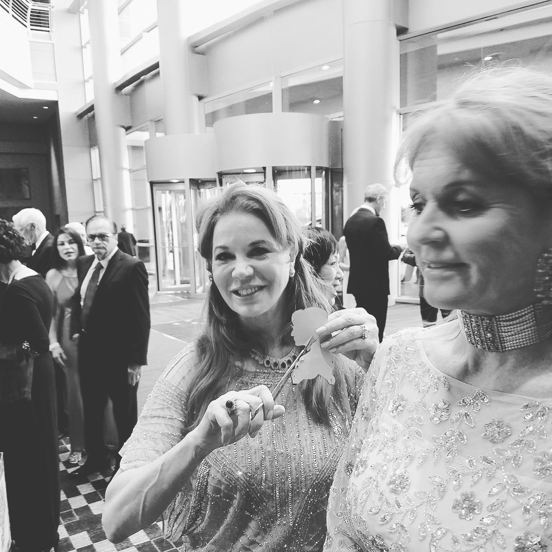 Two women at a formal event, one smiling and holding a spoon, the other looking away, with people in background.