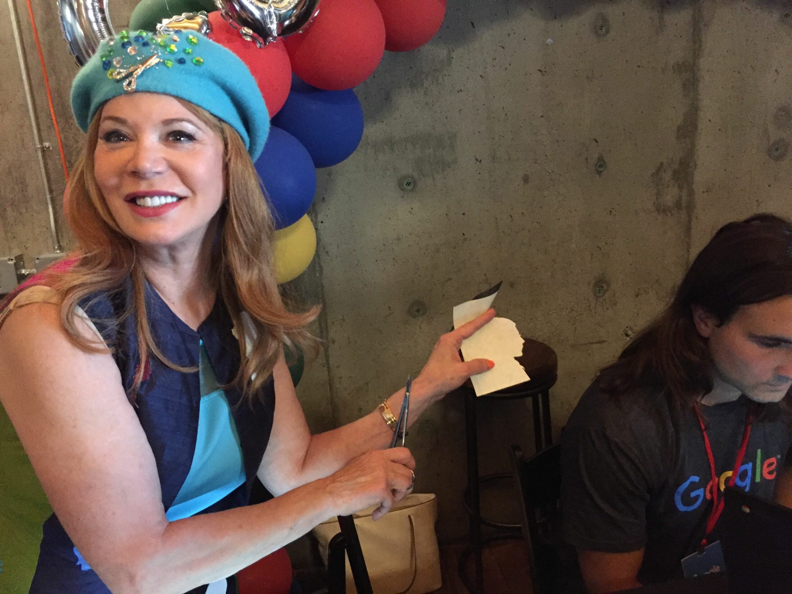 Woman smiling, wearing a blue hat with decorations, holding a piece of paper, with balloons in the background.