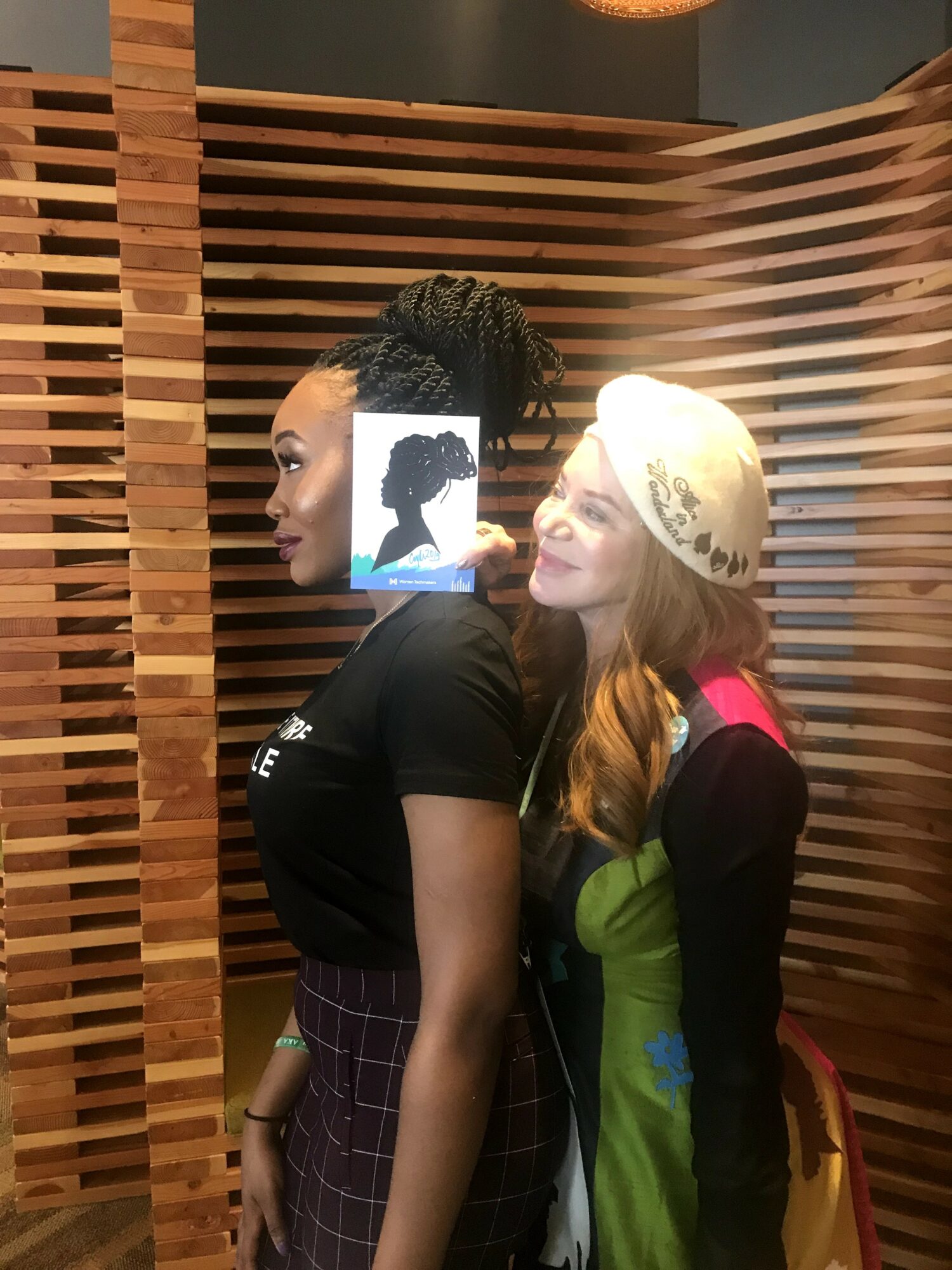 Two women stand close together, one with braided hair and the other with a hat, in front of a wooden backdrop.