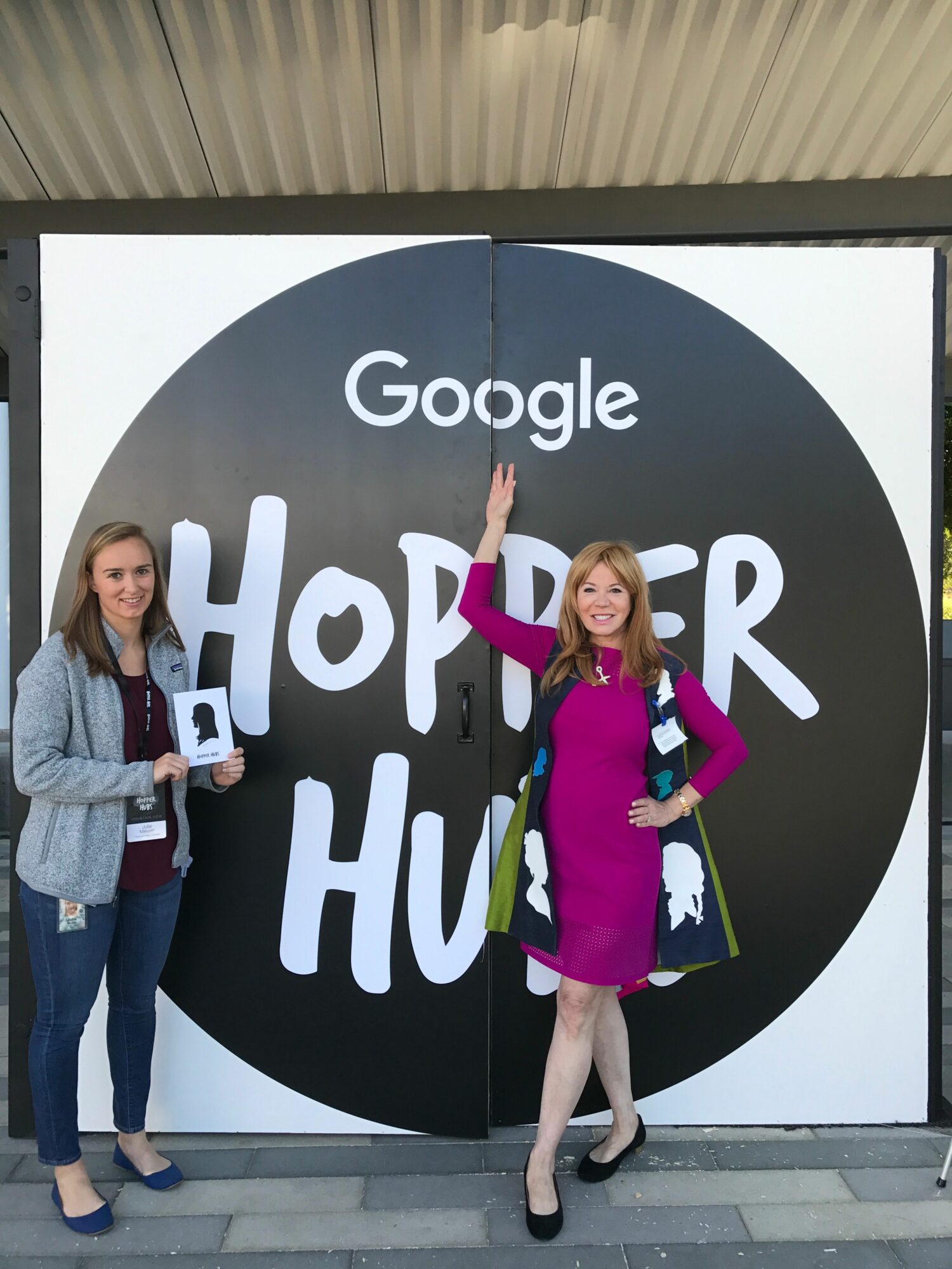 Two women standing in front of a large Google Hopper Hunt sign, one holding a phone, the other with hand raised.
