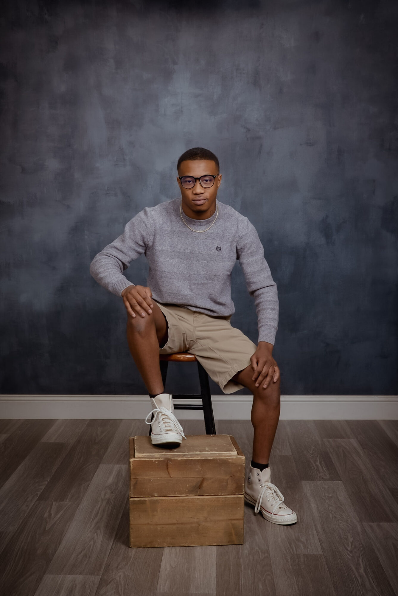 Young man with glasses in gray sweater and beige shorts, one foot on a wooden block, in a room with dark wall.