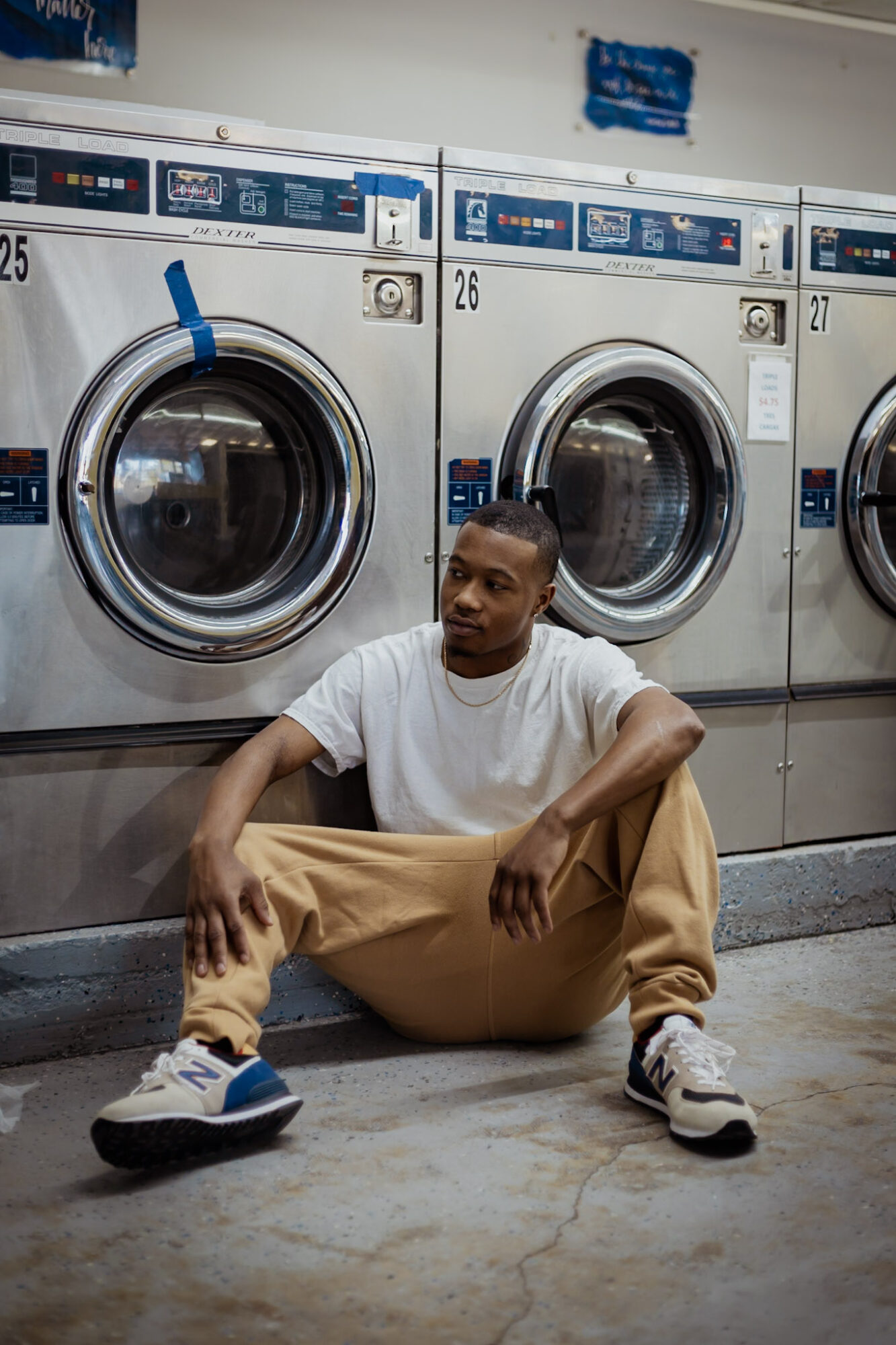 Young man sitting on the floor in front of washing machines, wearing a white t-shirt and tan pants.
