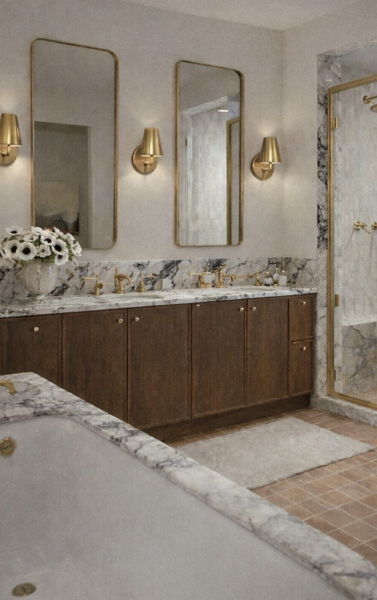 Bathroom with three mirrors, gold wall sconces, marble countertop, wooden cabinets, and a shower with marble and gold fixtures.