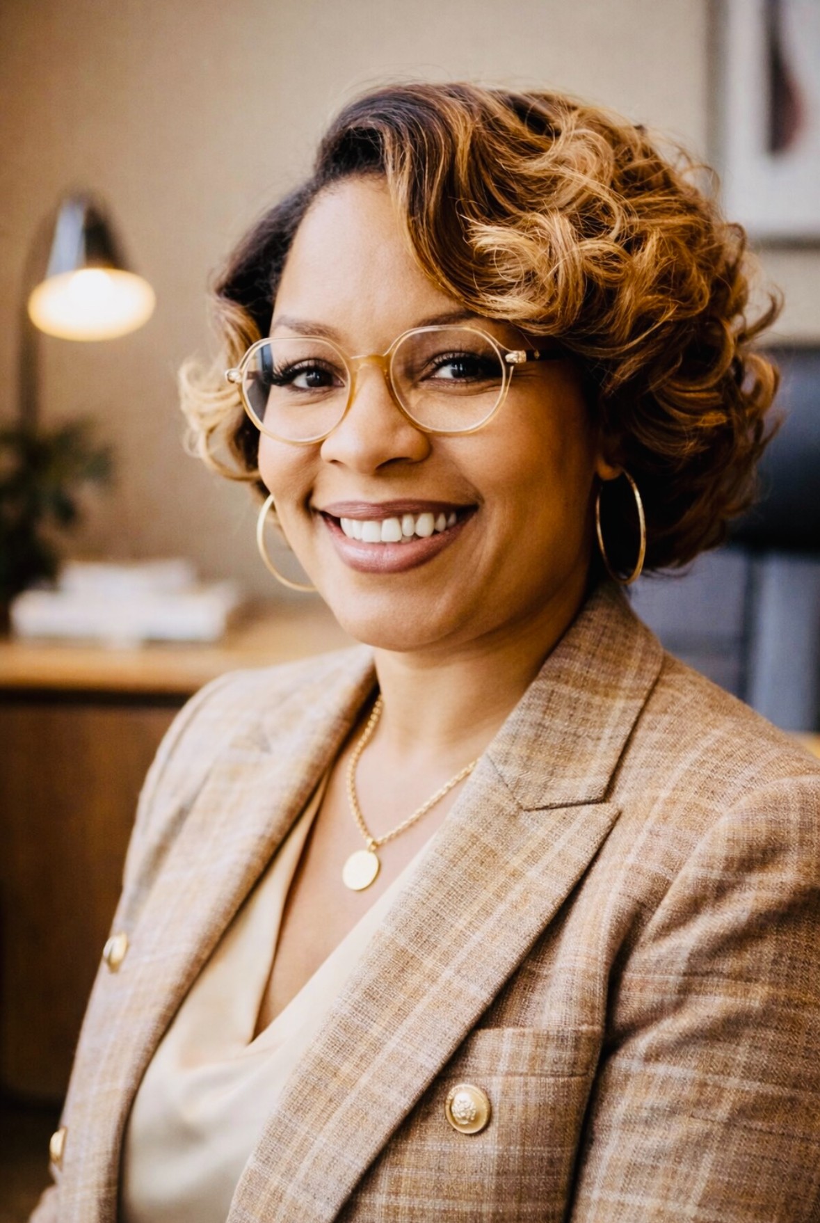 Woman with curly hair, glasses, and hoop earrings smiling in an indoor setting.