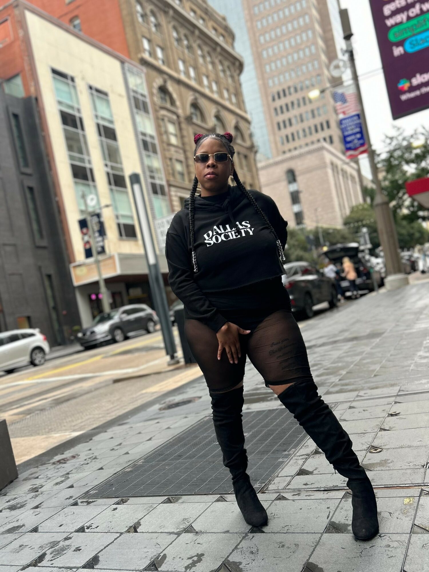 Person standing on city sidewalk wearing sunglasses, black hoodie, ripped jeans, and boots, with tall buildings in background.