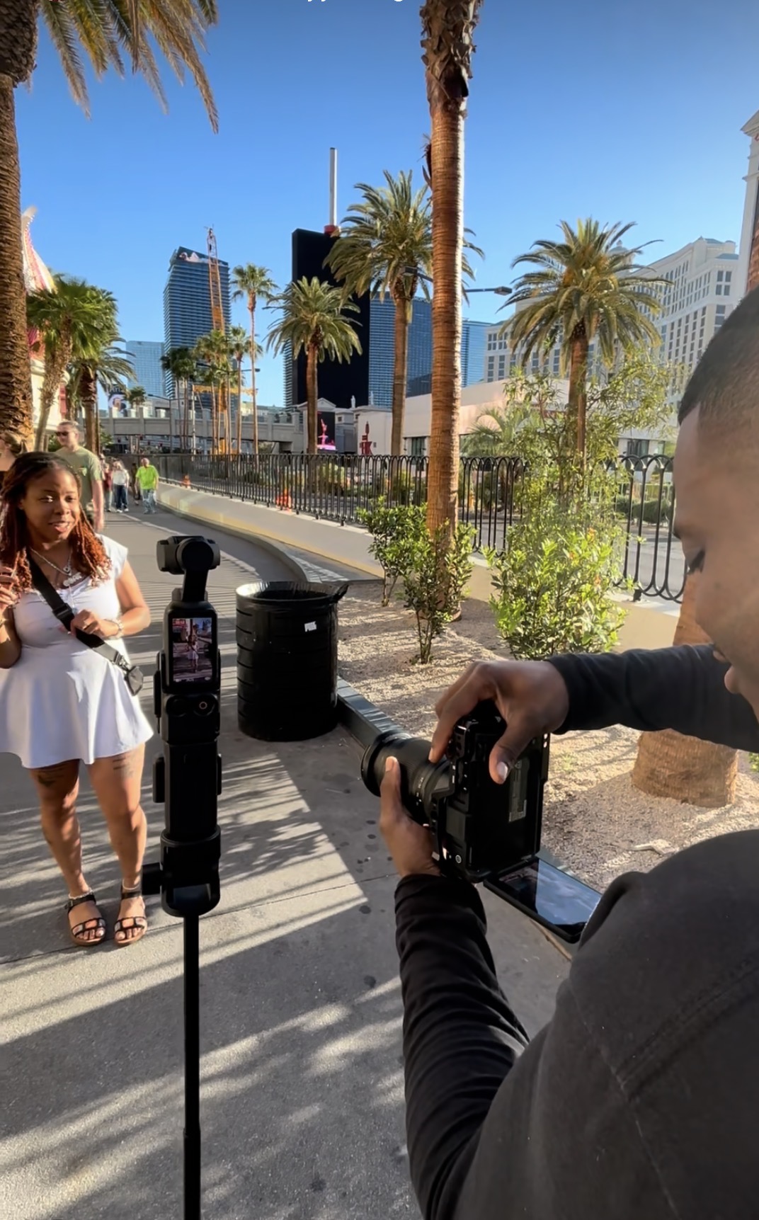Person taking a photo of a woman in a white dress outdoors with palm trees and city buildings in the background.
