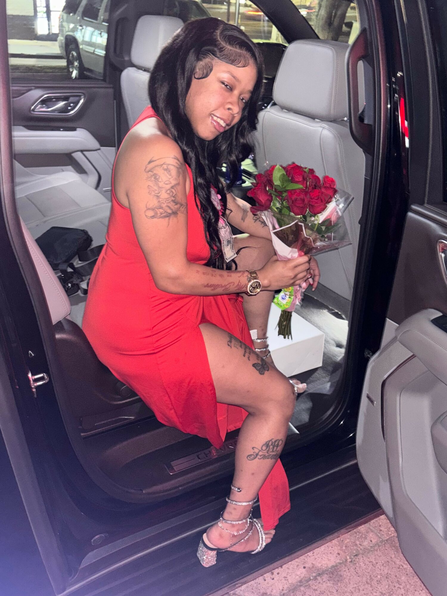 Woman in red dress sitting in car seat holding a bouquet of red roses, smiling.