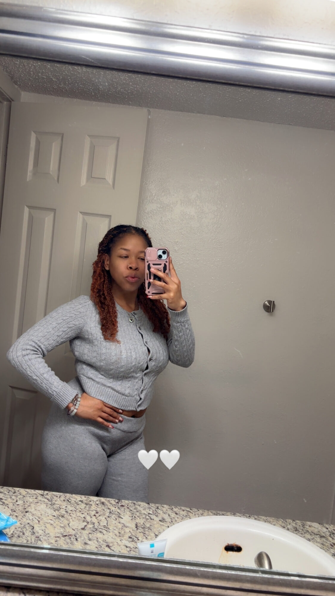 Woman taking mirror selfie in a bathroom, wearing gray outfit, with a door and wall in the background.