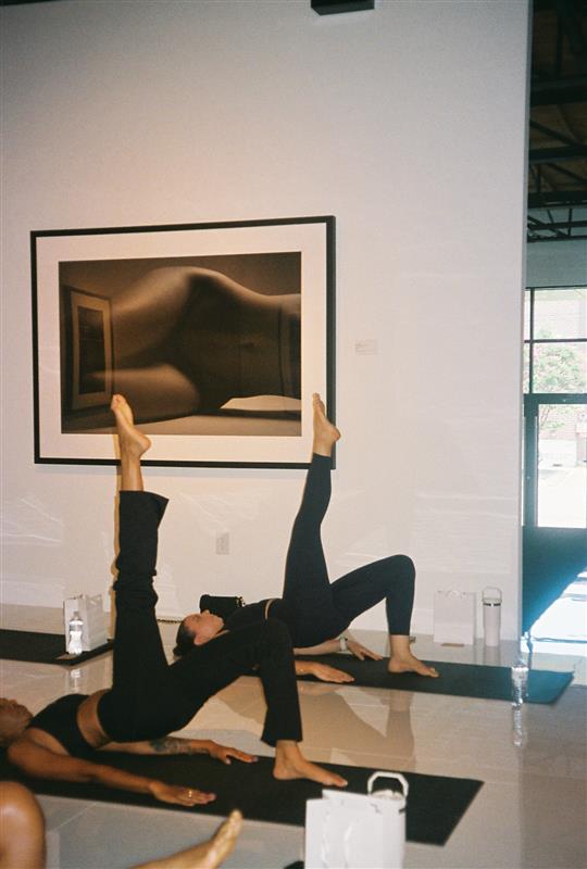 Two people perform yoga poses on mats in front of a framed photograph on a white wall, with water bottles nearby.