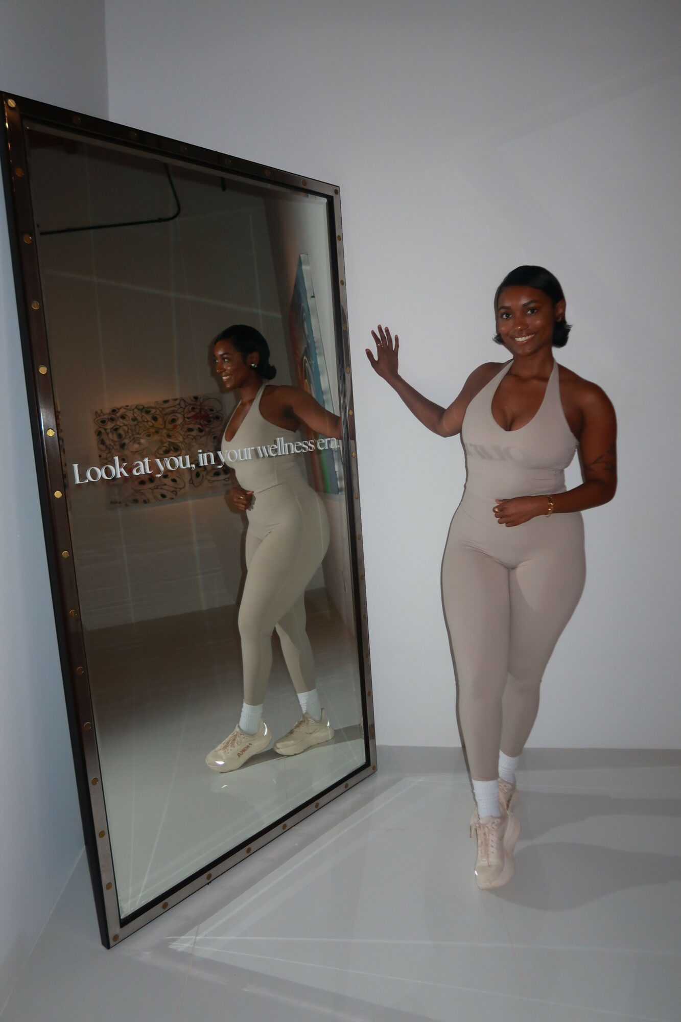 Woman standing next to a mirror, smiling, with her reflection visible, in a minimalistic room.
