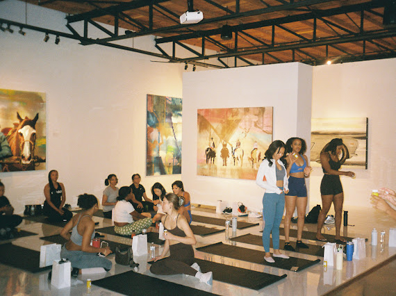 Group of people in an art gallery, some sitting on mats, others standing and talking, with colorful paintings on walls.