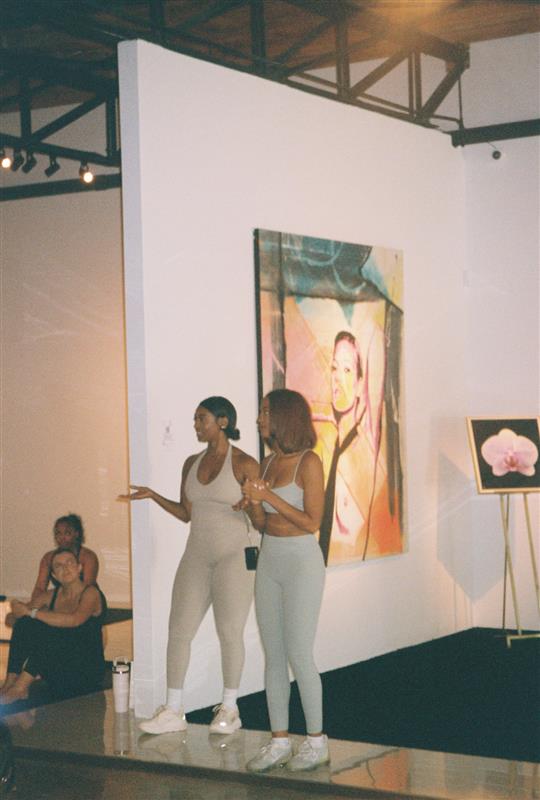 Three women in an art gallery, two standing and one sitting, with paintings on white walls behind them.