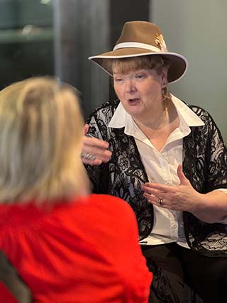 Older woman wearing a hat, white shirt, and black lace jacket, speaking to a person in red.
