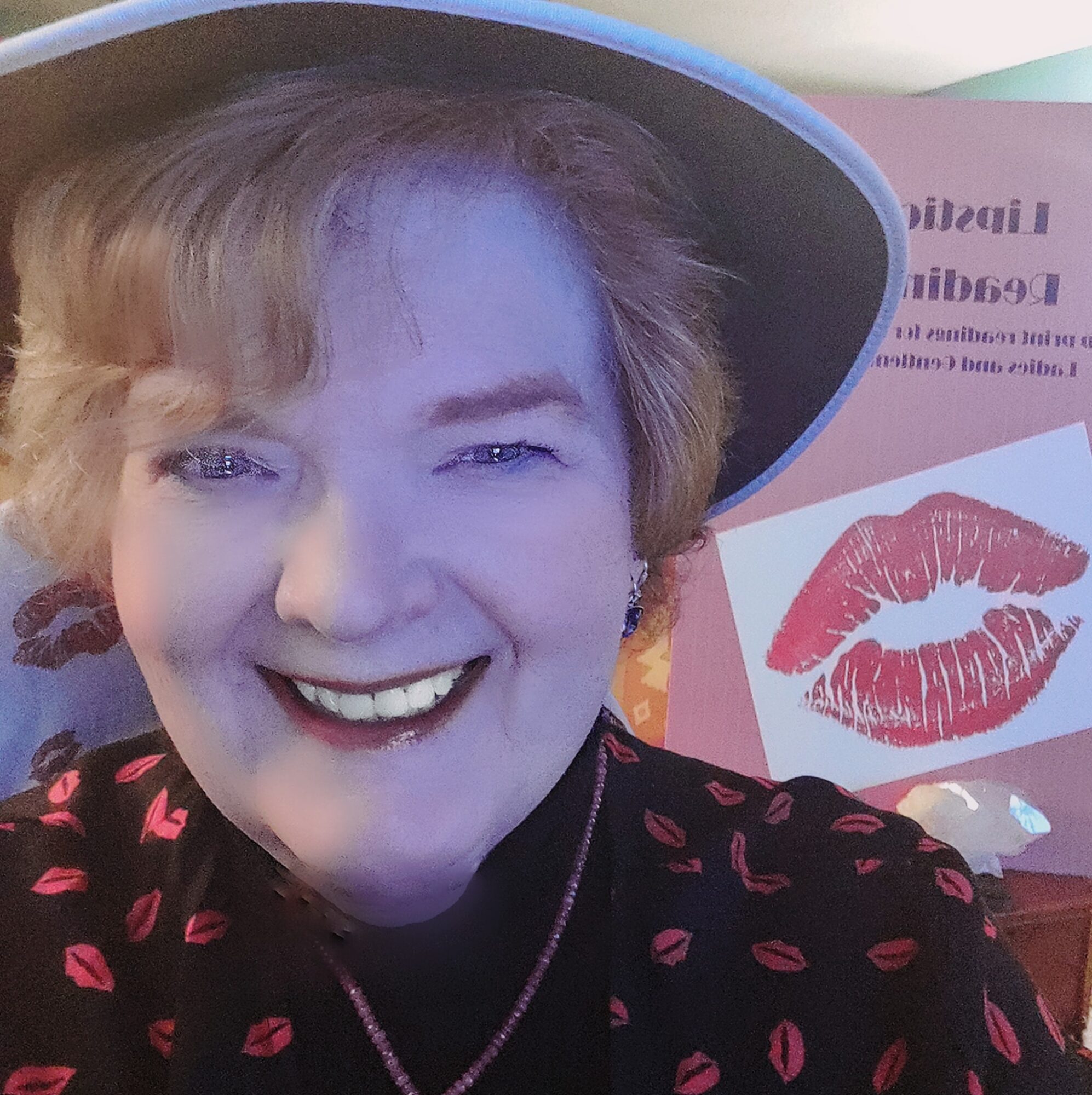 Smiling woman wearing a hat and a black top with pink lips pattern, in front of a poster with lipstick marks.