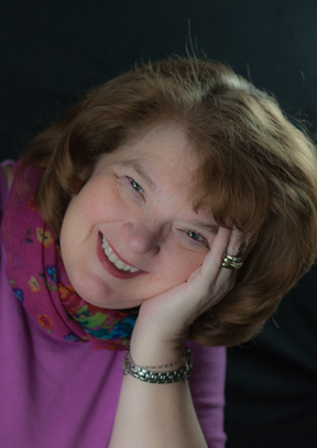 Smiling woman with red hair resting her head on her hand, wearing a colorful scarf and pink top.