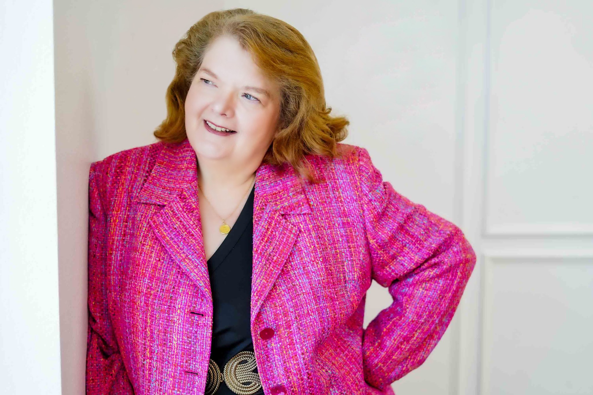 Woman with shoulder-length hair wearing a pink blazer, smiling, standing indoors.