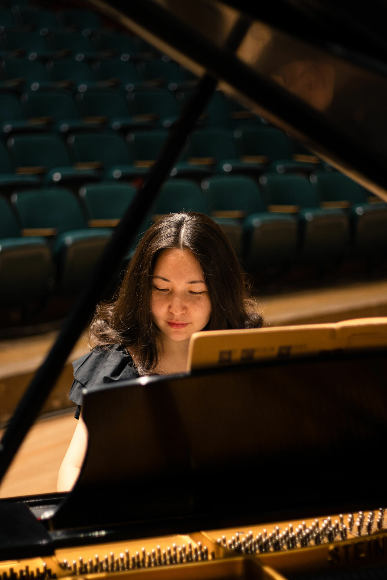 Young woman playing grand piano in an empty auditorium with blue seats.