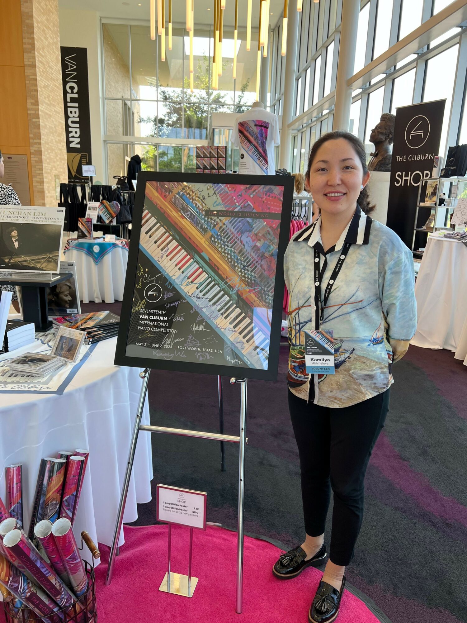 Woman standing next to a colorful poster at an indoor event, smiling, with tables and banners in background.